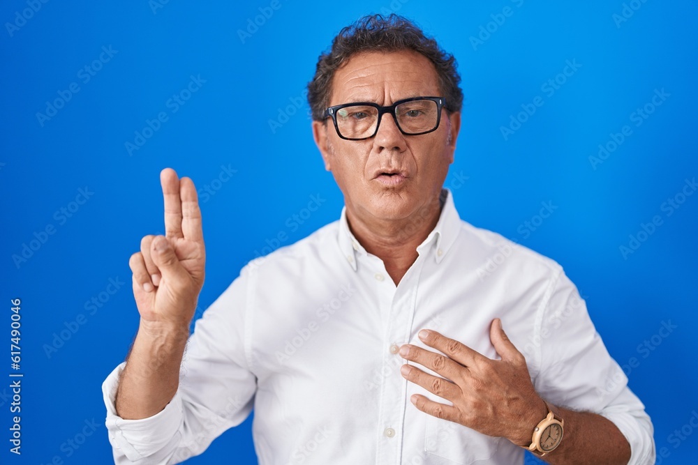 Middle age hispanic man standing over blue background smiling swearing with hand on chest and fingers up, making a loyalty promise oath Middle age hispanic man standing over blue background smiling swearing with hand on chest and fingers up, making a loyalty promise oath