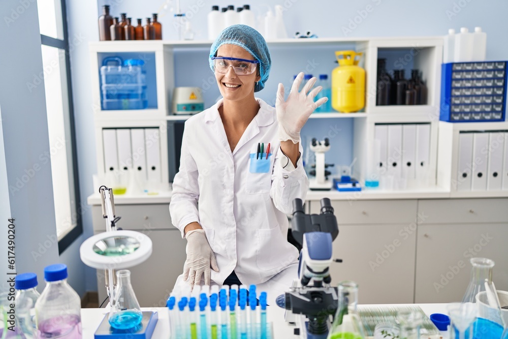 Fototapeta premium Brunette woman working at scientist laboratory waiving saying hello happy and smiling, friendly welcome gesture