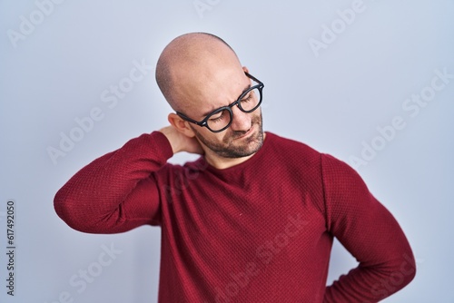 Young bald man with beard standing over white background wearing glasses suffering of neck ache injury, touching neck with hand, muscular pain