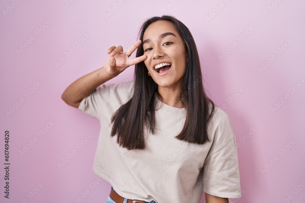 Young hispanic woman standing over pink background doing peace symbol with fingers over face, smiling cheerful showing victory Young hispanic woman standing over pink background doing peace symbol with fingers over face, smiling cheerful showing victory