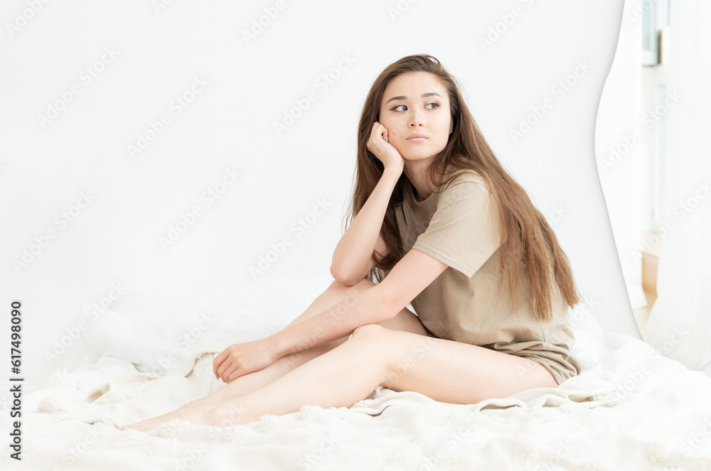 Naklejka premium Sad and lonely girl sits in bed on the white linen and looks out the window
