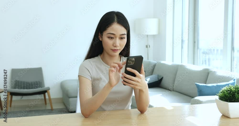 Asian woman sit at table in living room hold smartphone, spend time at home, use modern tech, enjoy remote chat communication with friends, text messages in social media, browse news using smartphone Asian woman sit at table in living room hold smartphone, spend time at home, use modern tech, enjoy remote chat communication with friends, text messages in social media, browse news using smartphone