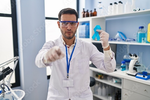 Young hispanic man with beard working at scientist laboratory holding blue ribbon pointing with finger to the camera and to you  confident gesture looking serious