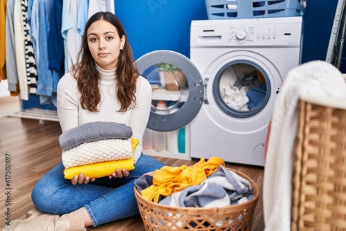 Young hispanic woman holding clean laundry relaxed with serious expression on face. simple and natural looking at the camera.
