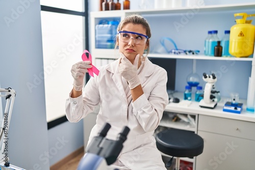 Young woman working at scientist laboratory holding pink ribbon serious face thinking about question with hand on chin, thoughtful about confusing idea