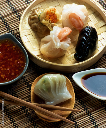 Assorted Dim Sum in Bamboo Steamed Bowl and Hagao with Shrimp on Wooden Plate with Red Chili and Soy Sauces and Chopsticks closeup on Straw Mat background photo