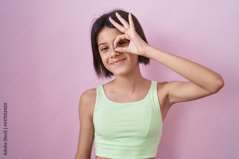 Young girl standing over pink background doing ok gesture with hand smiling, eye looking through fingers with happy face. Young girl standing over pink background doing ok gesture with hand smiling, eye looking through fingers with happy face.
