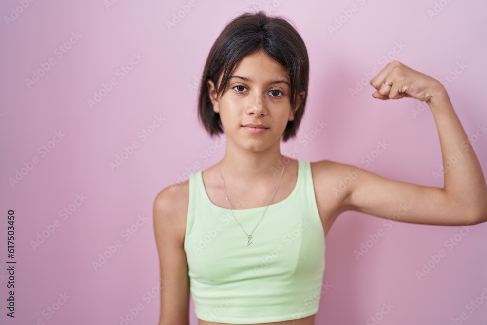 Young girl standing over pink background strong person showing arm muscle, confident and proud of power Young girl standing over pink background strong person showing arm muscle, confident and proud of power