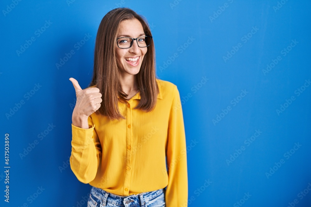 Young woman wearing glasses standing over blue background doing happy thumbs up gesture with hand. approving expression looking at the camera showing success. Young woman wearing glasses standing over blue background doing happy thumbs up gesture with hand. approving expression looking at the camera showing success.