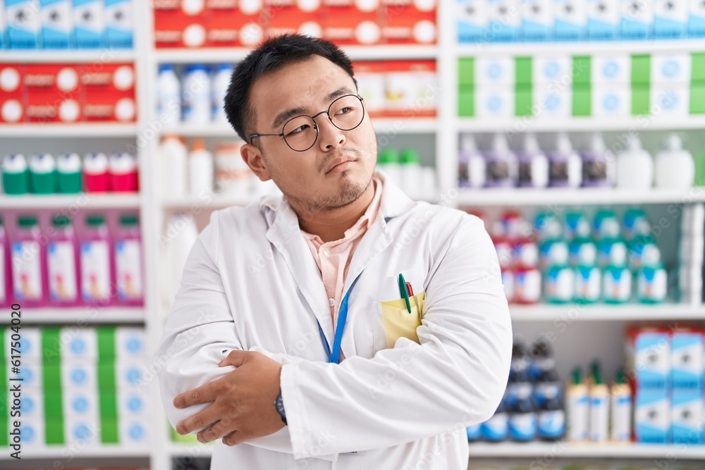 Chinese young man working at pharmacy drugstore looking to the side with arms crossed convinced and confident Chinese young man working at pharmacy drugstore looking to the side with arms crossed convinced and confident