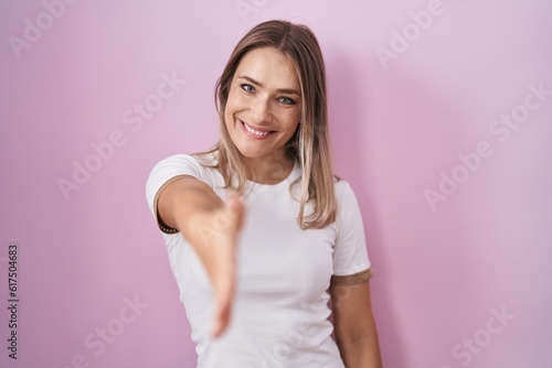 Blonde caucasian woman standing over pink background smiling friendly offering handshake as greeting and welcoming. successful business.