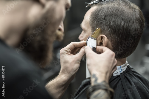 Unapproachable barber with a beard and a tattoo is cutting the hair of his client in the barbershop. He is using a cutting comb and a hair clipper. Customer has hairgrips on the head. Closeup.