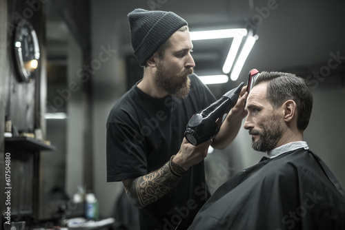 Unequaled barber with a beard and a tattoo is drying the hair of his client in a black cutting hair cape in the barbershop. He is using a black hairdryer and a hairbrush. Horizontal.