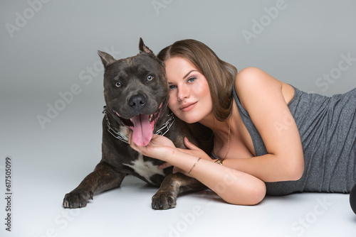 Beautiful sporty young woman lying on floor and hugging adult grey amstafford terrier dog. Studio shot over gray background. Copy space.