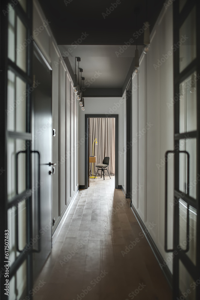 Low lighted corridor in a modern style with white walls and a parquet on the floor. There is an entrance door to a room with a wooden table, black chair and a trendy yellow lamp. Vertical.