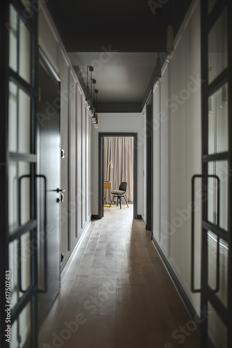 Low lighted corridor in a modern style with white walls and a parquet on the floor. There is an entrance door to a room with a wooden table, black chair and a trendy yellow lamp. Vertical. © Designpics