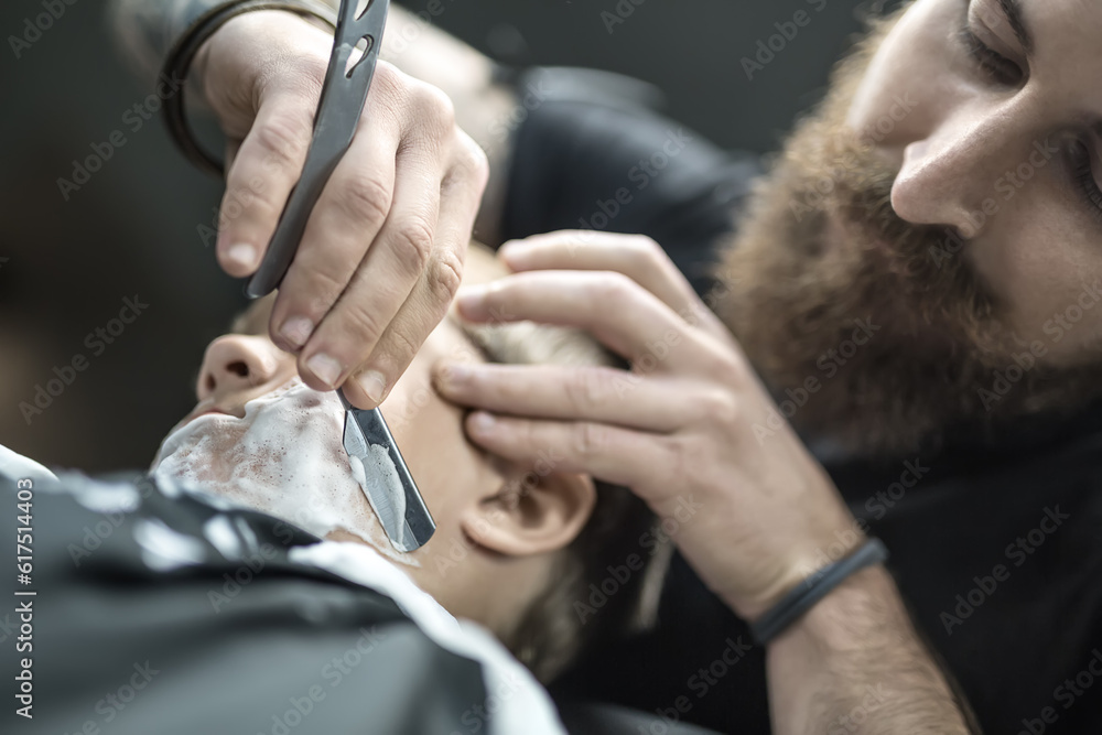 Obraz premium Macro photo of the little boy with a shaving foam on his face in the barbershop. Barber is shaving kid's face with the help of the straight razor. Low aperture photo. Horizontal.