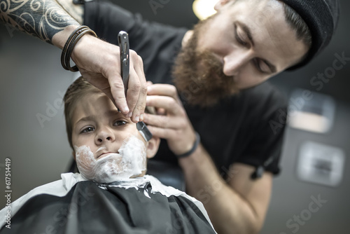 Small kid with a shaving foam on the face in the barbershop. He wears a black salon cape. Bearded barber with a tattoo is shaving boy's face with the help of the straight razor. Low aperture photo.