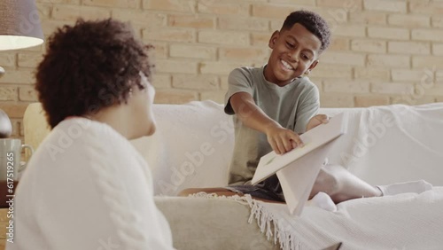 menino Brasileiro sentado no sofa da sala com seu caderno pedindo ajuda de sua mae para fazer a tarefa da escola sentada no chao da sala trabalhando photo