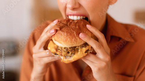 Hungry caucasian woman enjoy eating tasty hamburger, biting home-prepared or delivered junk food meal, closeup, cropped