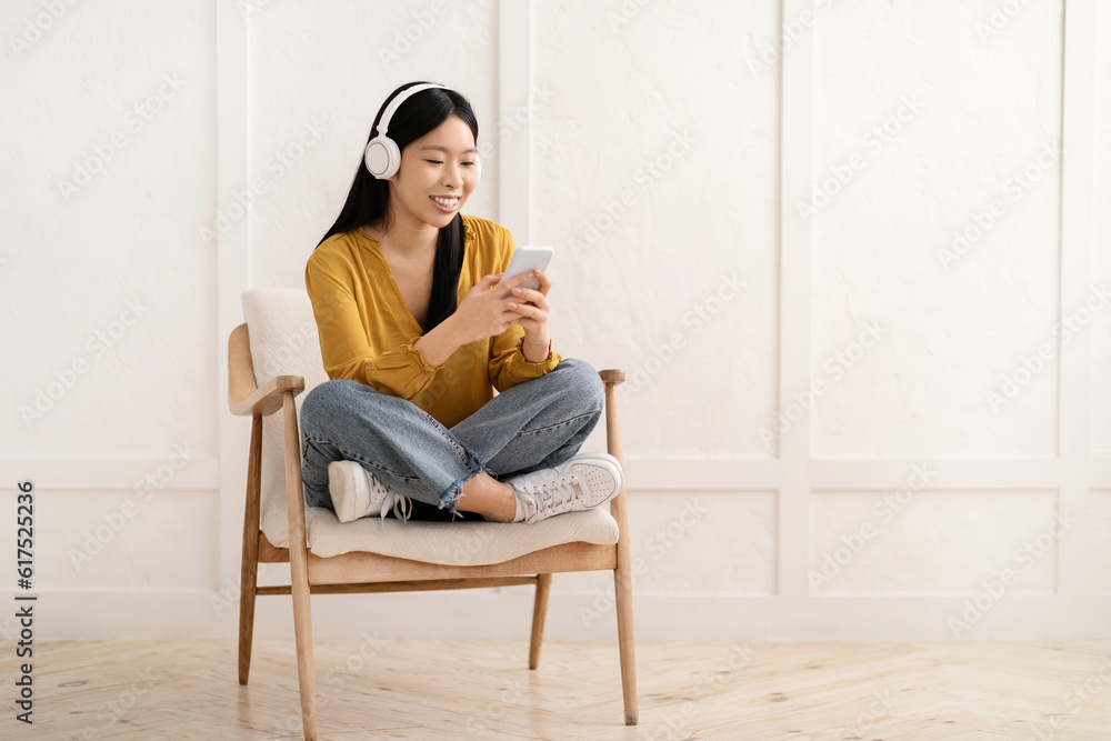 Cheerful asian woman sitting in armchair, using cell phone, headphones Cheerful asian woman sitting in armchair, using cell phone, headphones