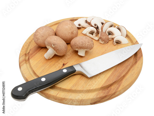 Sharp knife with whole chestnut mushrooms and slices on a circular wooden chopping board, isolated on a white background