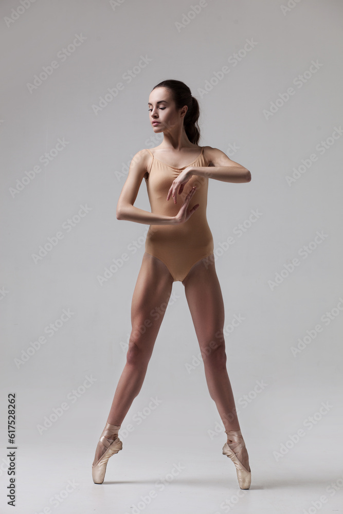 Fototapeta premium young beautiful ballet dancer in beige swimsuit posing on pointes on light grey studio background