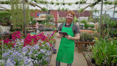 A male staff of Flower Shop holding tablet device while standing inside local business smiling at camera. Young redhair man employee wearing apron and using modern technology