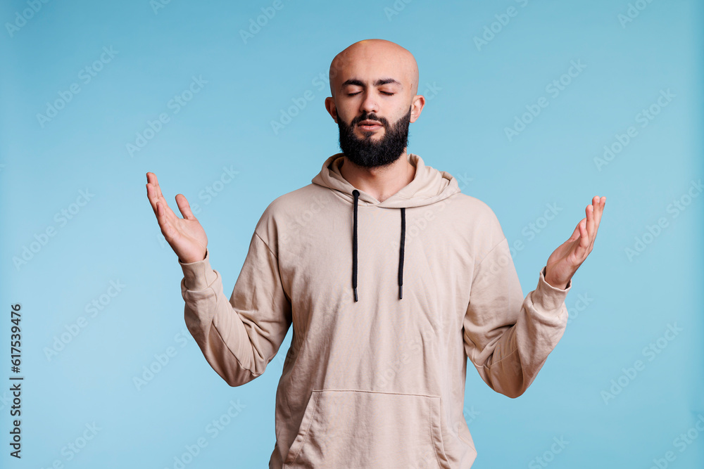 Fototapeta premium Arab man praying and making worship gesture with open arms and closed eyes. Young religious person praising, pleading to god with raised hands and asking for forgiveness concept
