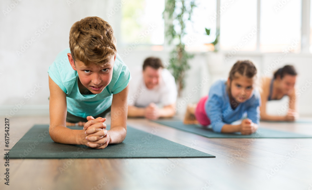 Diligent preteen boy practicing plank pose of yoga on black mat together with his parents and sister