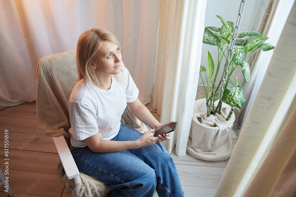 Young blonde woman using cell phone at home. Girl is resting, chating and looking in camera on smartphone near window at home. Nice atmosphere. The concept of a cozy home and communication by Internet Young blonde woman using cell phone at home. Girl is resting, chating and looking in camera on smartphone near window at home. Nice atmosphere. The concept of a cozy home and communication by Internet