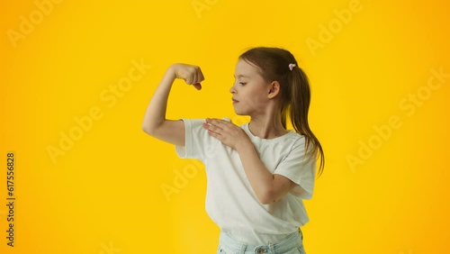 Young girl bending her arm, demonstrate her bicep and touch it on yellow background. Concept of proper child development, healthy nutrition and physical training