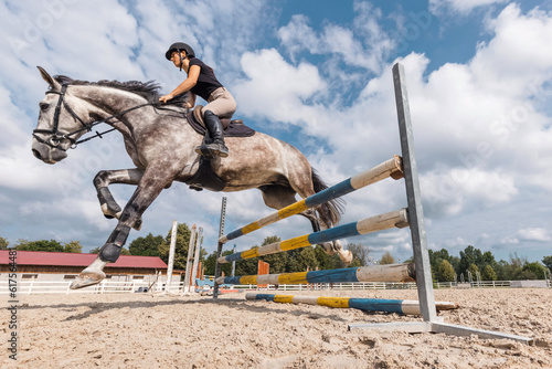 Girl rider on a dapple gray horse jumping over a hurdle at the manege of equestrian center on a sunny day © 24K-Production