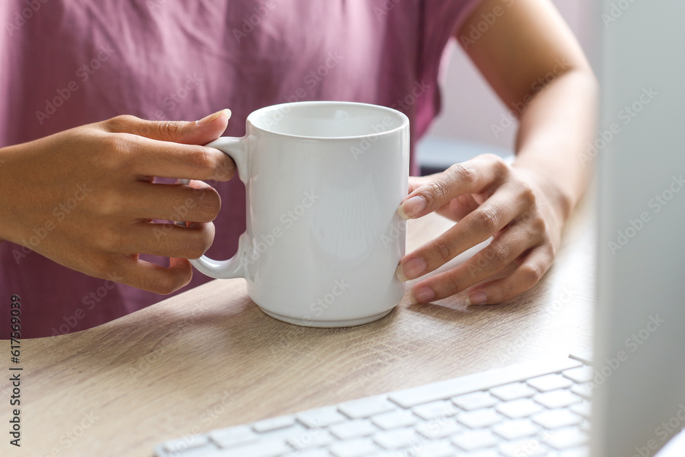 Woman sitting on desk and looking at screen computer while holding white mug mockup that can put logo, text or brand Woman sitting on desk and looking at screen computer while holding white mug mockup that can put logo, text or brand