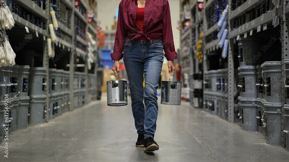 Woman shown from waist down, carrying to cans of paint walking down paint aisle in hardware store