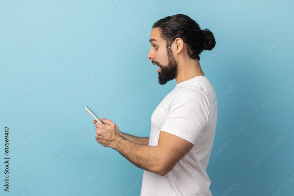 Side view of surprised man with beard wearing white T-shirt expressing shock while using cell phone, searching web, astonishing news. Indoor studio shot isolated on blue background. Side view of surprised man with beard wearing white T-shirt expressing shock while using cell phone, searching web, astonishing news. Indoor studio shot isolated on blue background.