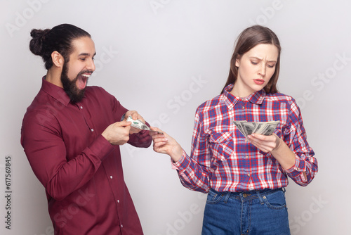 Portrait of serious rich confident woman and man gigolo standing together, wife giving money to her husband with indifferent face. Indoor studio shot isolated on gray background.