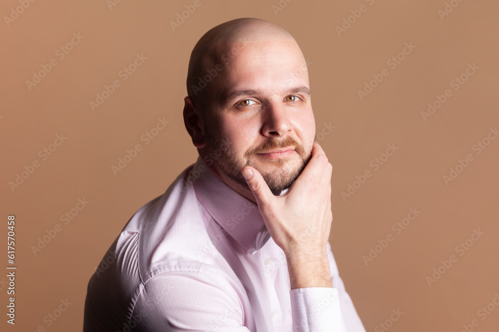 Side view portrait of cute positive smiling bald bearded man sitting holding chin, looking at camera with positive expression, wearing light pink shirt. Indoor studio shot isolated on brown background Side view portrait of cute positive smiling bald bearded man sitting holding chin, looking at camera with positive expression, wearing light pink shirt. Indoor studio shot isolated on brown background