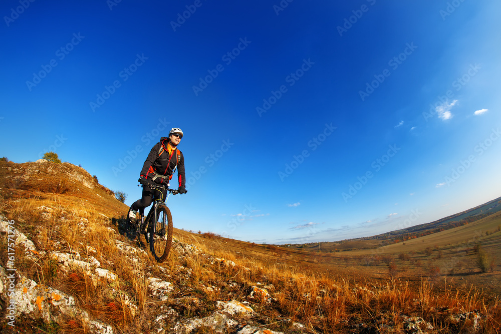 Wide angle view of a cyclist riding a bike on a nature trail in the mountains. people living a healthy lifestyle. Cyclist with helmet and gjasses. Beautiful landscape with hill and blue sky. Spring se