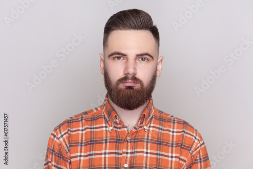 Portrait of good-looking serious bearded man standing looking at camera with strict bossy facial expression, wearing checkered shirt. Indoor studio shot isolated on gray background.