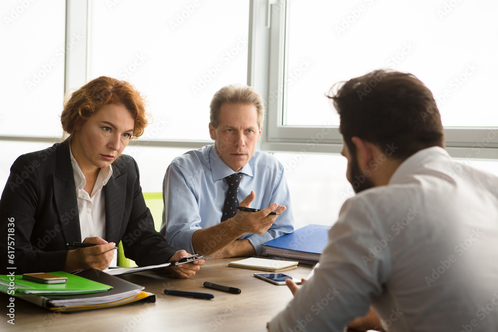 Business people taking interview while sitting at table in board room all together. Serious business workers listening to new worker sitting in front. Employee concept. Job concept.