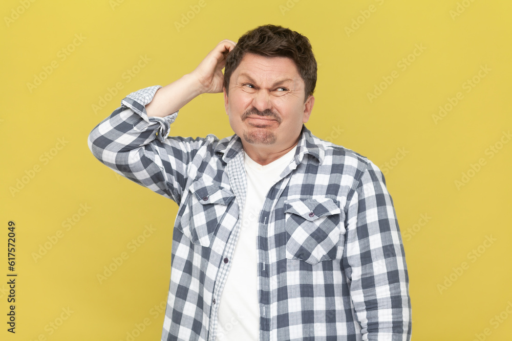 Portrait of thoughtful attractive middle aged man in casual checkered shirt, keeps hand on his head, thinking, trying to remember something. Indoor studio shot isolated on yellow background. Portrait of thoughtful attractive middle aged man in casual checkered shirt, keeps hand on his head, thinking, trying to remember something. Indoor studio shot isolated on yellow background.