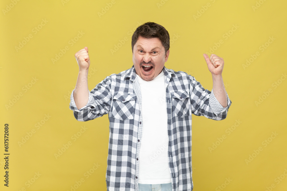 Extremely happy overjoyed middle aged man in casual checkered shirt standing with raised arms and clenched fists, winning lottery, celebrating. Indoor studio shot isolated on yellow background. Extremely happy overjoyed middle aged man in casual checkered shirt standing with raised arms and clenched fists, winning lottery, celebrating. Indoor studio shot isolated on yellow background.