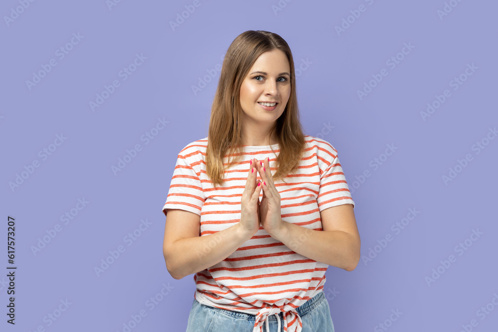 Cunning blond woman wearing striped T-shirt gesticulating and thinking over devious sly plan of revenge, scheming and conspiring villain plan. Indoor studio shot isolated on purple background.