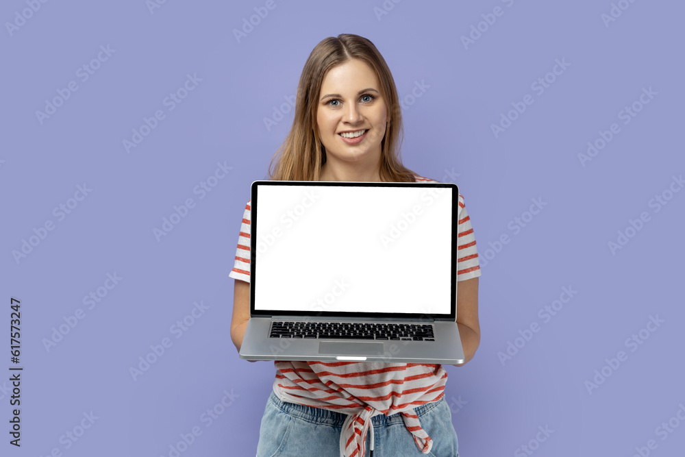 Portrait of happy joyful blond woman wearing striped T-shirt holding laptop with white empty display with empty space for advertisement. Indoor studio shot isolated on purple background. Portrait of happy joyful blond woman wearing striped T-shirt holding laptop with white empty display with empty space for advertisement. Indoor studio shot isolated on purple background.