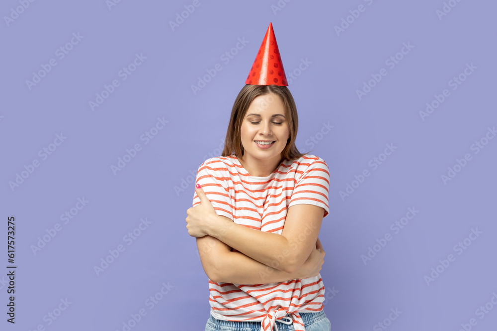Portrait of self-satisfied egoistic woman in party cone celebrating her birthday, embracing herself and smiling with pleasure, feeling self-pride. Indoor studio shot isolated on purple background. Portrait of self-satisfied egoistic woman in party cone celebrating her birthday, embracing herself and smiling with pleasure, feeling self-pride. Indoor studio shot isolated on purple background.