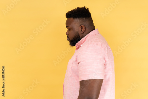 Side view of depressed sad young adult man wearing pink shirt being upset of bad news, looking ahead with frowning face, expressing sadness. Indoor studio shot isolated on yellow background. photo