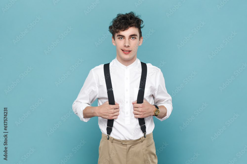 Portrait of smiling satisfied young man wearing white shirt holding his suspenders, looking at camera with positive facial expression. Indoor studio shot isolated on blue background. Portrait of smiling satisfied young man wearing white shirt holding his suspenders, looking at camera with positive facial expression. Indoor studio shot isolated on blue background.