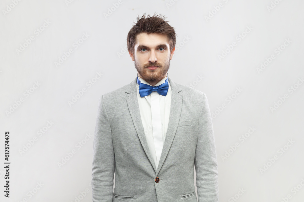 Portrait of calm positive bearded man standing looking at camera, having relaxed facial expression, wearing grey suit and blue bow tie. Indoor studio shot isolated on gray background. Portrait of calm positive bearded man standing looking at camera, having relaxed facial expression, wearing grey suit and blue bow tie. Indoor studio shot isolated on gray background.