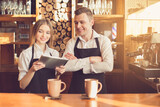 Professional barista. Young woman and man in aprons smiling and standing at bar counter. Woman using tablet computer